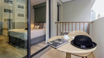 Un balcón con una cama, una mesa con un sombrero y un libro, y vistas a edificios.