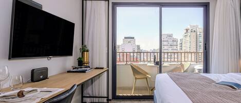 Quarto de hotel moderno com uma cama grande, uma escrivaninha com televisão de tela plana, um espaço de refeições com mesa e cadeiras, e uma varanda com vista para a cidade.