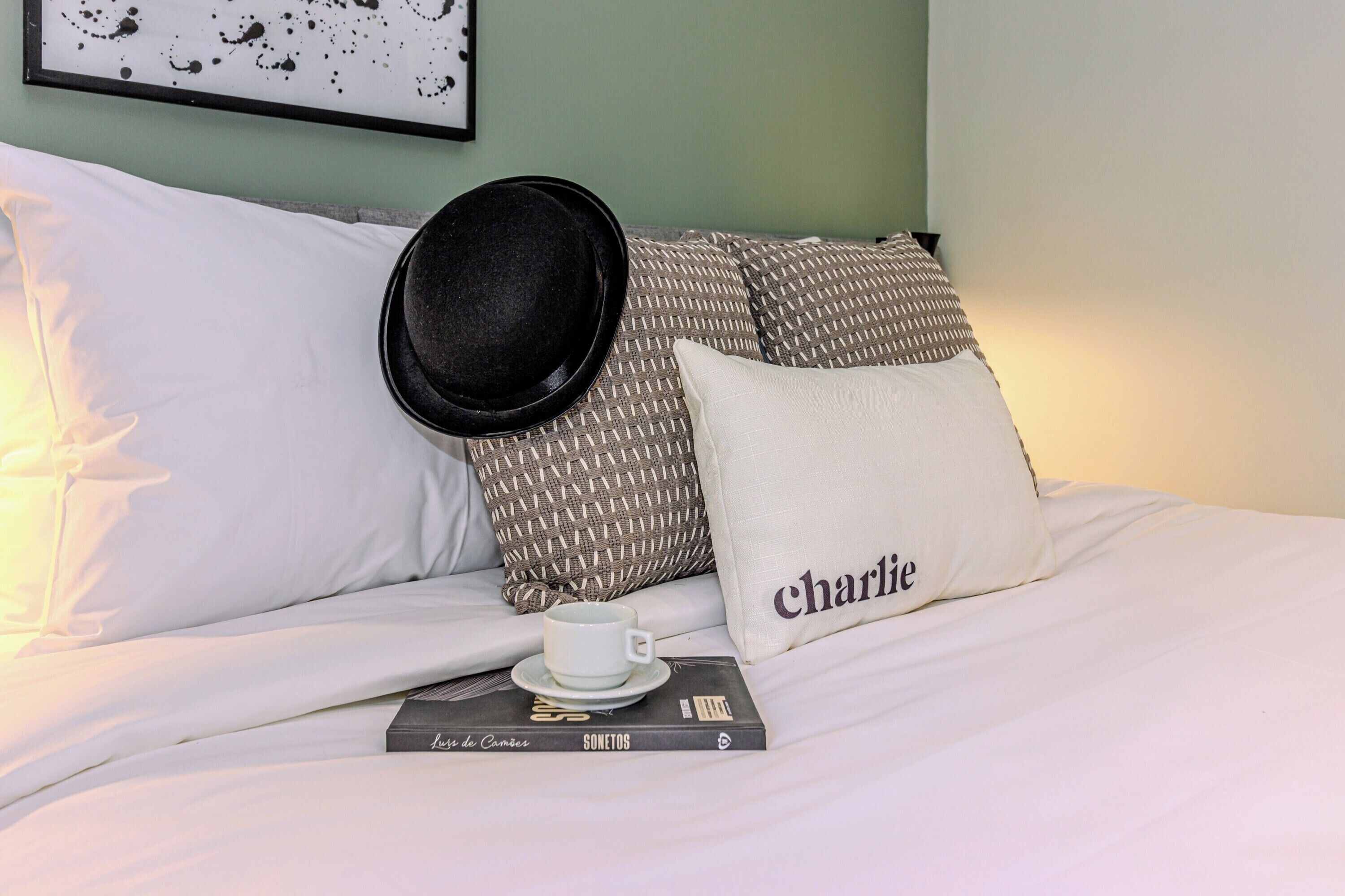 A neatly made bed with white linens, a black hat, a pillow with the name 'charlie', and a book titled 'Sometos'.