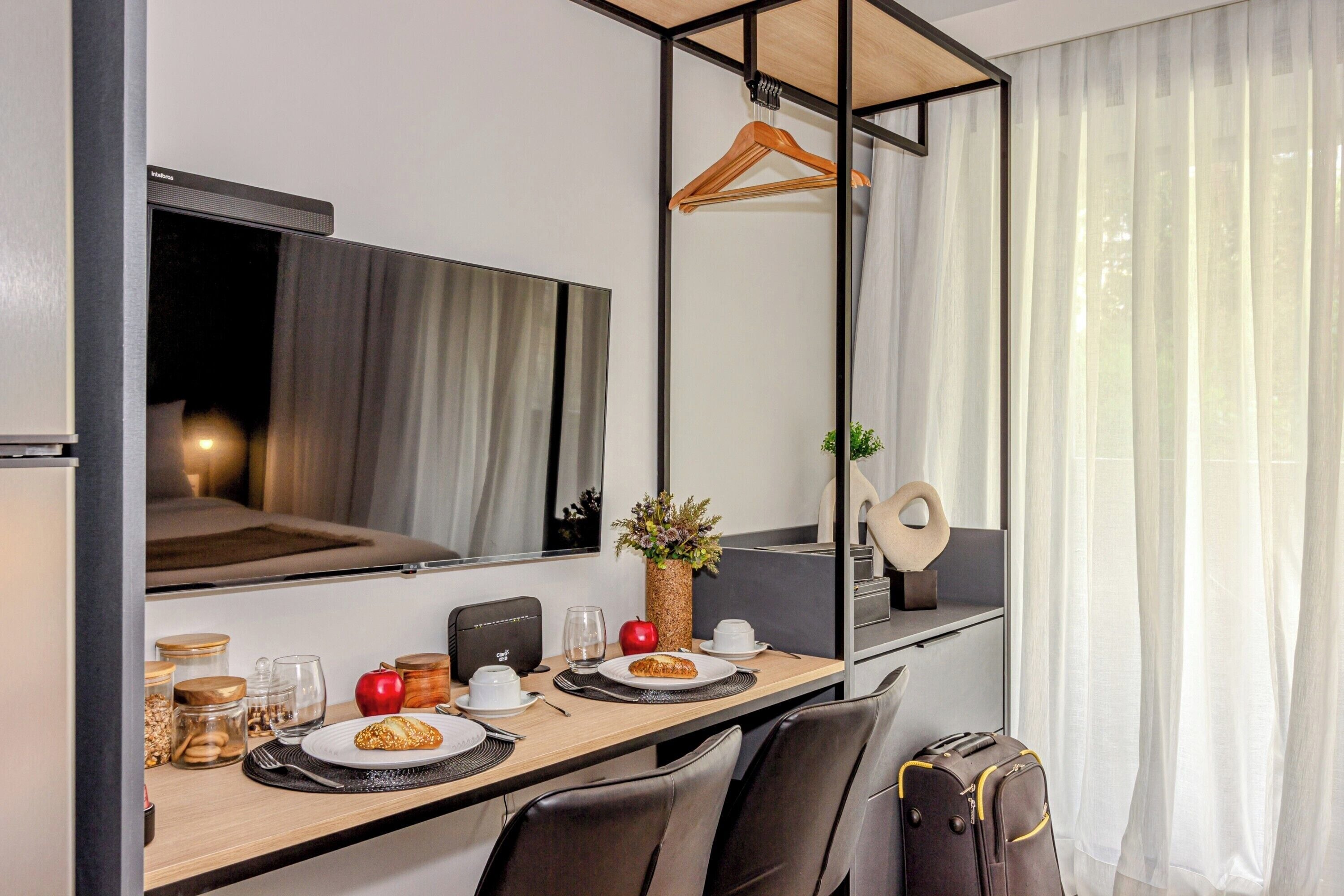 A hotel room with a dining table set for breakfast, a mirror, a hanging coat rack, and a suitcase on the floor.