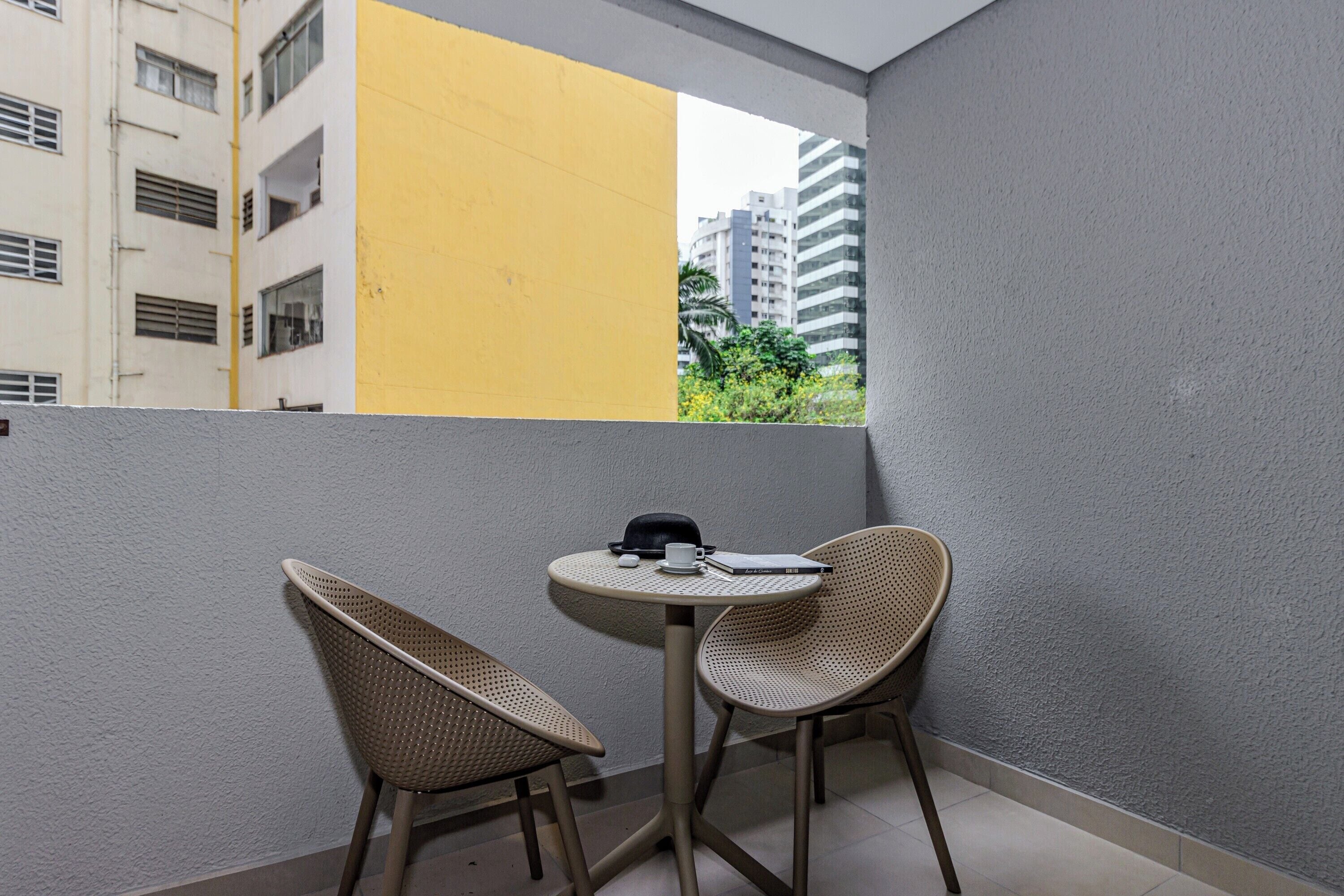 Ein Balkon mit zwei Korbstühlen, einem kleinen runden Tisch und Blick auf ein gelbes Gebäude sowie weitere Bauten.