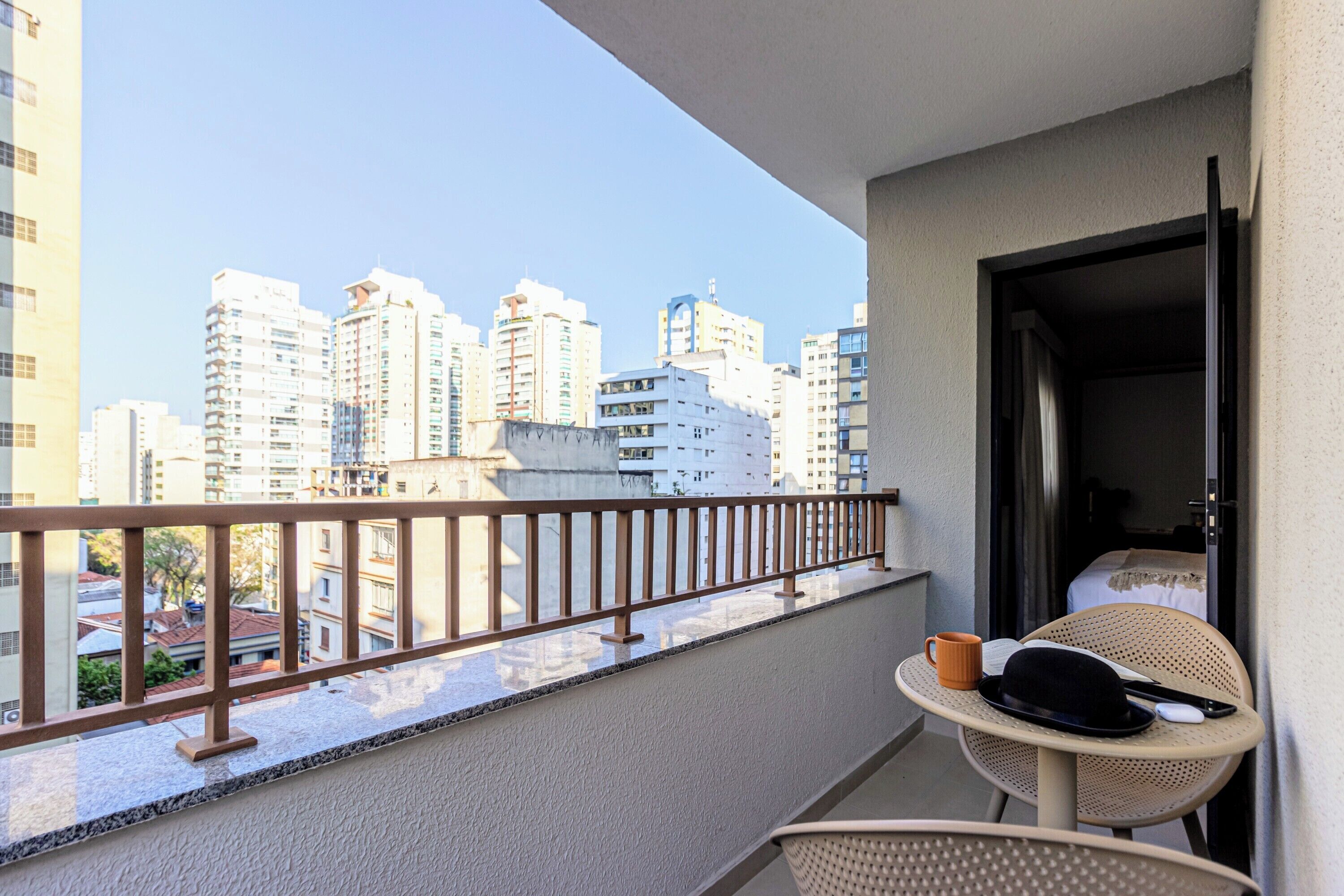 A balcony with a view of tall buildings, a table with a hat and a mug, and a chair.