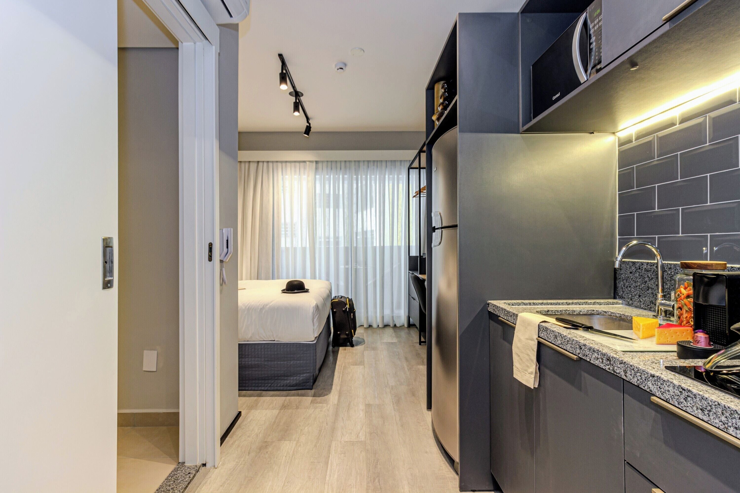 A modern hotel room with a bed, a desk, and a kitchenette.