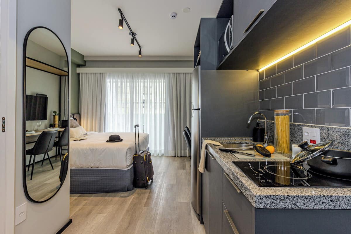 Une chambre d’hôtel compacte comprenant un lit, un bureau, une chaise, une valise, une kitchenette avec une cuisinière et un grand miroir.