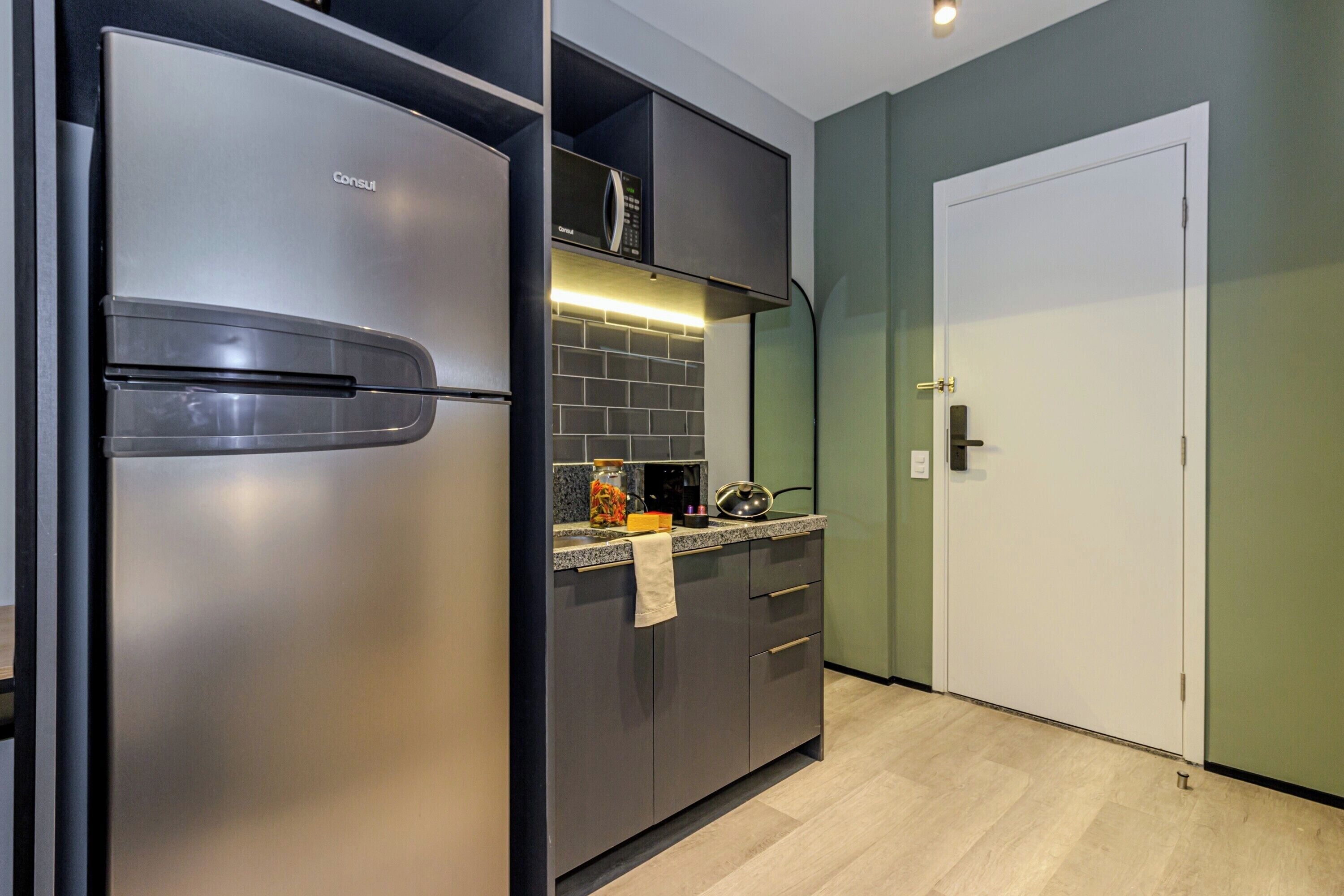 A modern kitchen with a stainless steel refrigerator, a microwave, and a built-in oven. There is a small countertop with a kettle and a jar. The kitchen has dark cabinets and a light-colored floor.