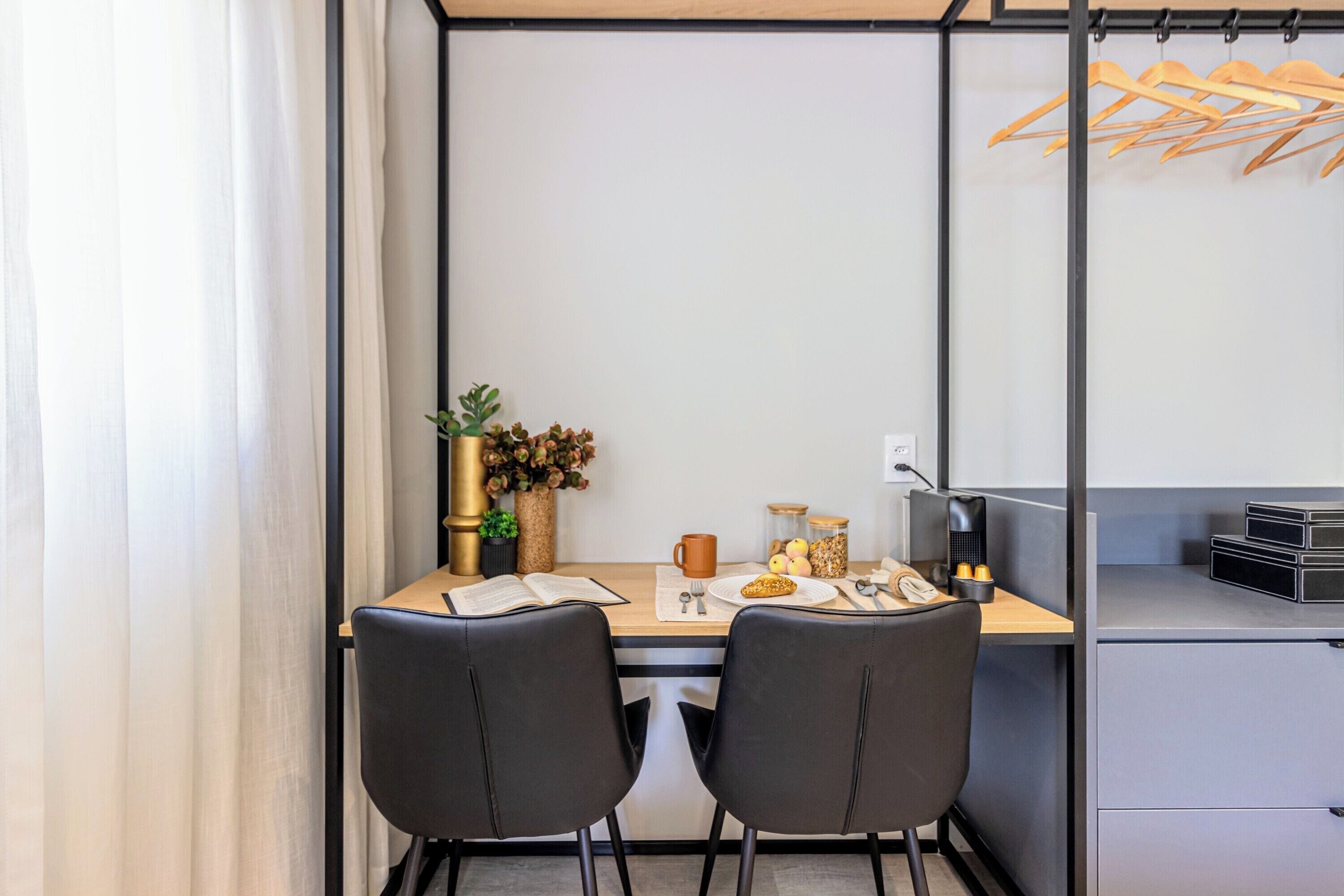 A modern dining area with a wooden table, two black chairs, a vase with flowers, a coffee machine, and a hanging wooden rack.