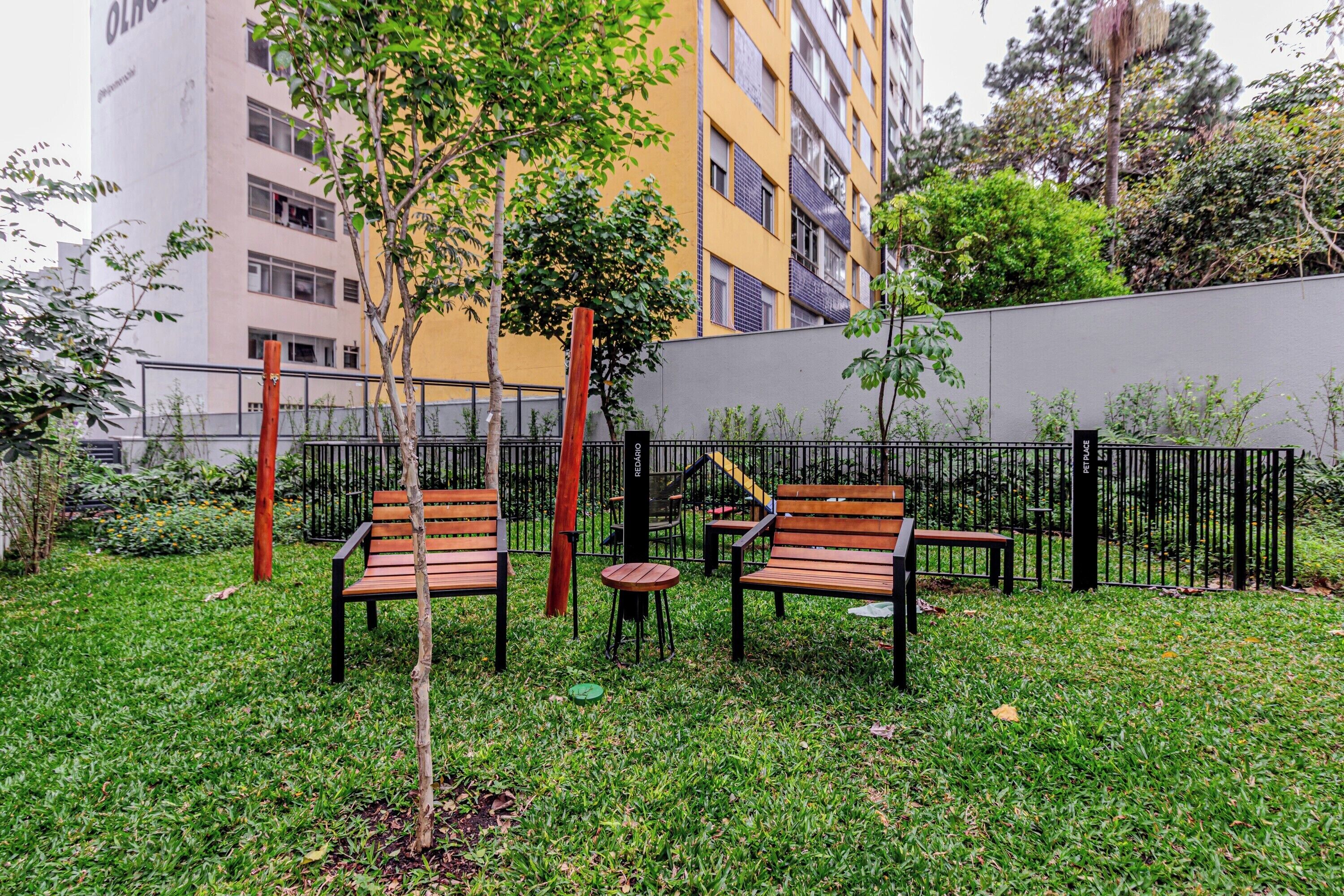 Un área de estar al aire libre con bancos y una pequeña mesa, rodeada por una cerca baja y follaje.