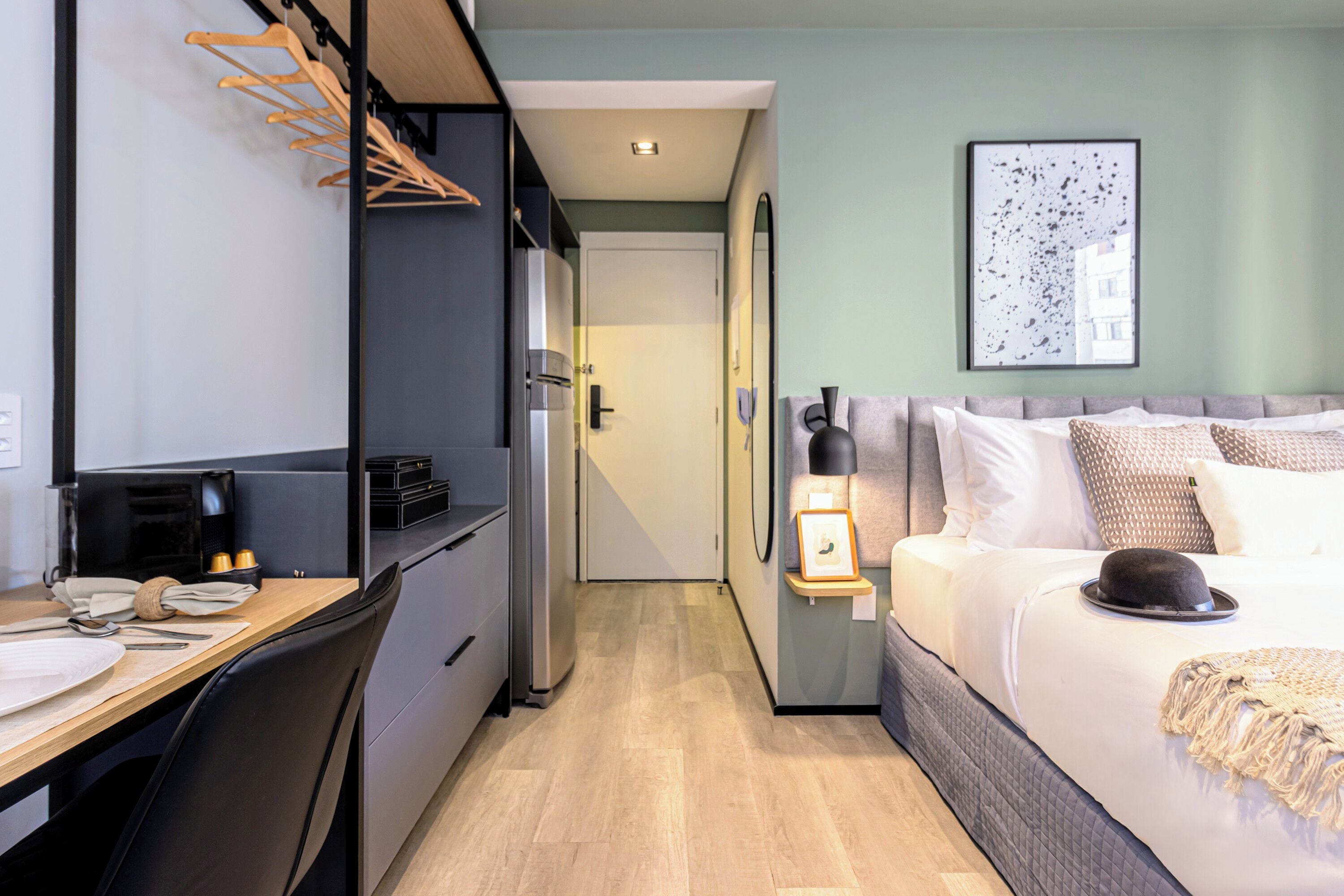 A compact hotel room with a kitchenette, a bed with a decorative hat, and a framed picture on the wall.