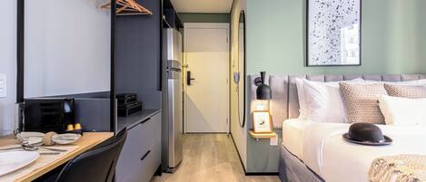A compact hotel room with a kitchenette, a bed with a decorative hat, and a framed picture on the wall.