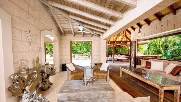 A spacious living area with a high ceiling, wooden beams, a large window with a view of greenery, and a cozy seating arrangement. - Kyalami - by Zenbreak (Holetown)
