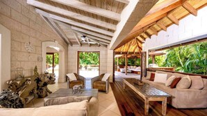 A spacious living area with a high ceiling, wooden beams, and a view of lush greenery outside. - Kyalami - by Zenbreak (Holetown)