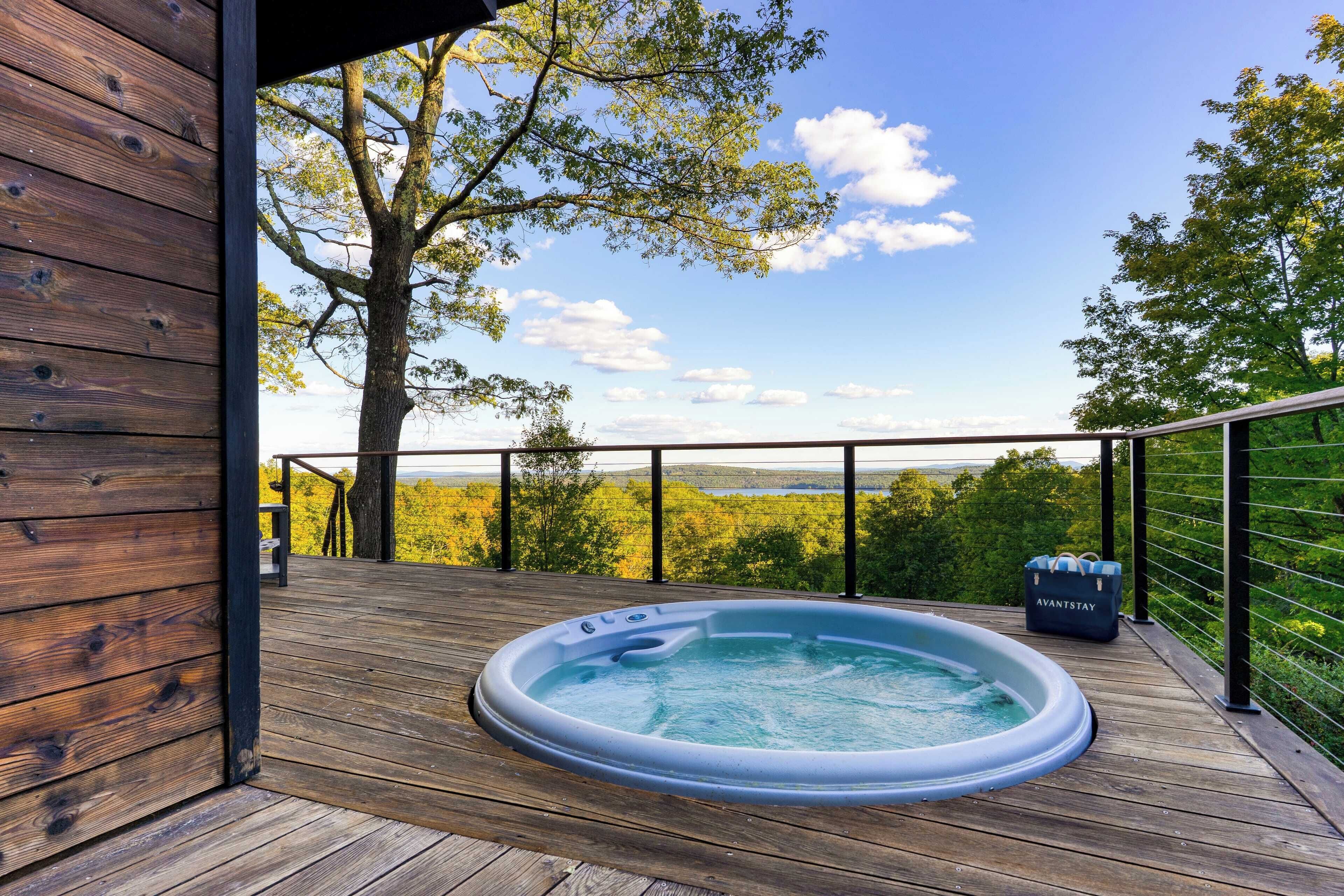 A wooden deck with a hot tub and a suitcase labeled "AVANTSTAY" on a balcony overlooking a forested landscape.