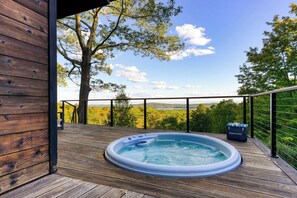 A wooden deck with a hot tub and a suitcase labeled "AVANTSTAY" on a balcony overlooking a forested landscape. - Modern Catskills Escape w/ Open Living, Steam Shower, & Hot Tub w/ Mountain Views, Minutes From Woodstock & Trails Suncliff Vista (Glenford)