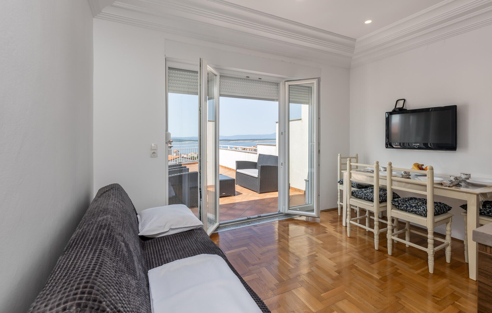 A room with a sofa, a dining table with chairs, a TV, and a view of the sea through a glass door.