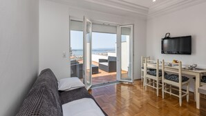 Ett rum med en soffa, ett matbord med stolar, en tv och utsikt över havet genom en glasdörr.