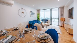 A modern dining area with a wooden table set for a meal, a blue sofa, and a large window offering a city view. - Sea-View 3BR, Rooftop Pool, Secure Parking & Study (Wynnum)