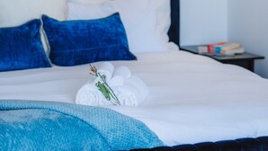 A neatly made bed with white linens and blue pillows, a small bedside table with books, and a wall-mounted picture of a sailboat. - Sea-View 3BR, Rooftop Pool, Secure Parking & Study (Wynnum)