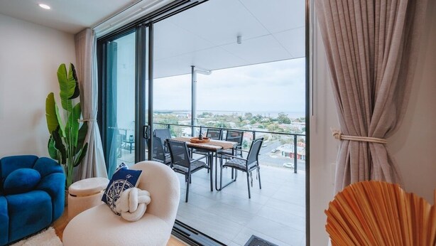 A modern living room with a large window, a dining area, and a view of the cityscape. - Sea-View 3BR, Rooftop Pool, Secure Parking & Study (Wynnum)