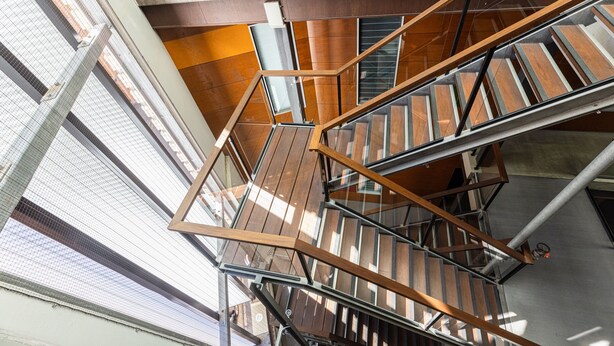 Un escalier intérieur à plusieurs niveaux, doté de rampes en métal et de marches en bois, entouré de murs de verre et de poutres métalliques.