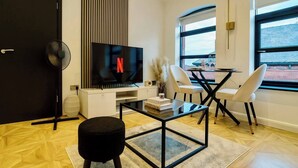 A modern living room with a flat-screen TV, a dining table with chairs, a black ottoman, and a large window with a view.