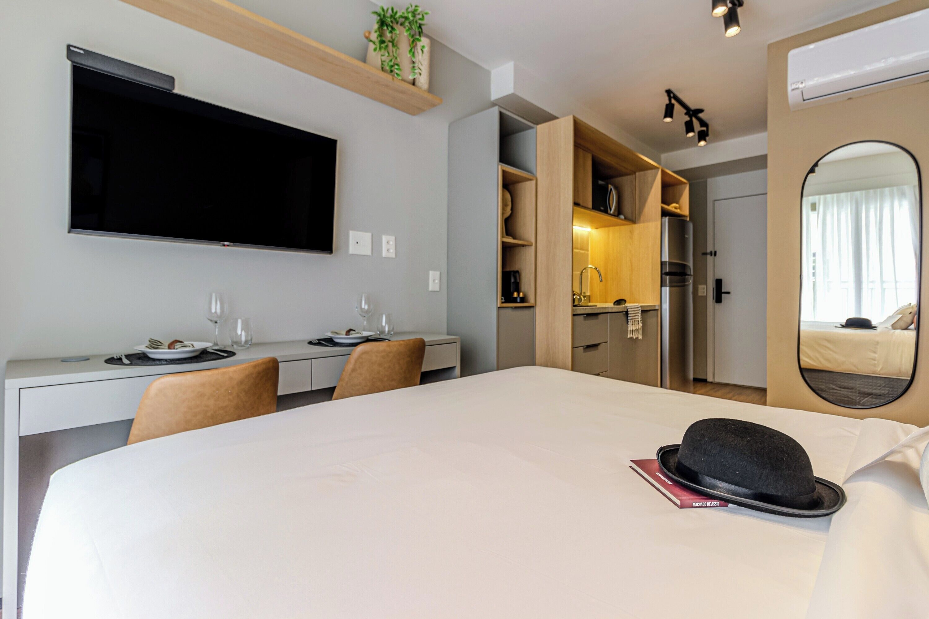 A hotel room with a bed, a desk, a flat-screen TV, a mirror, and a kitchenette.
