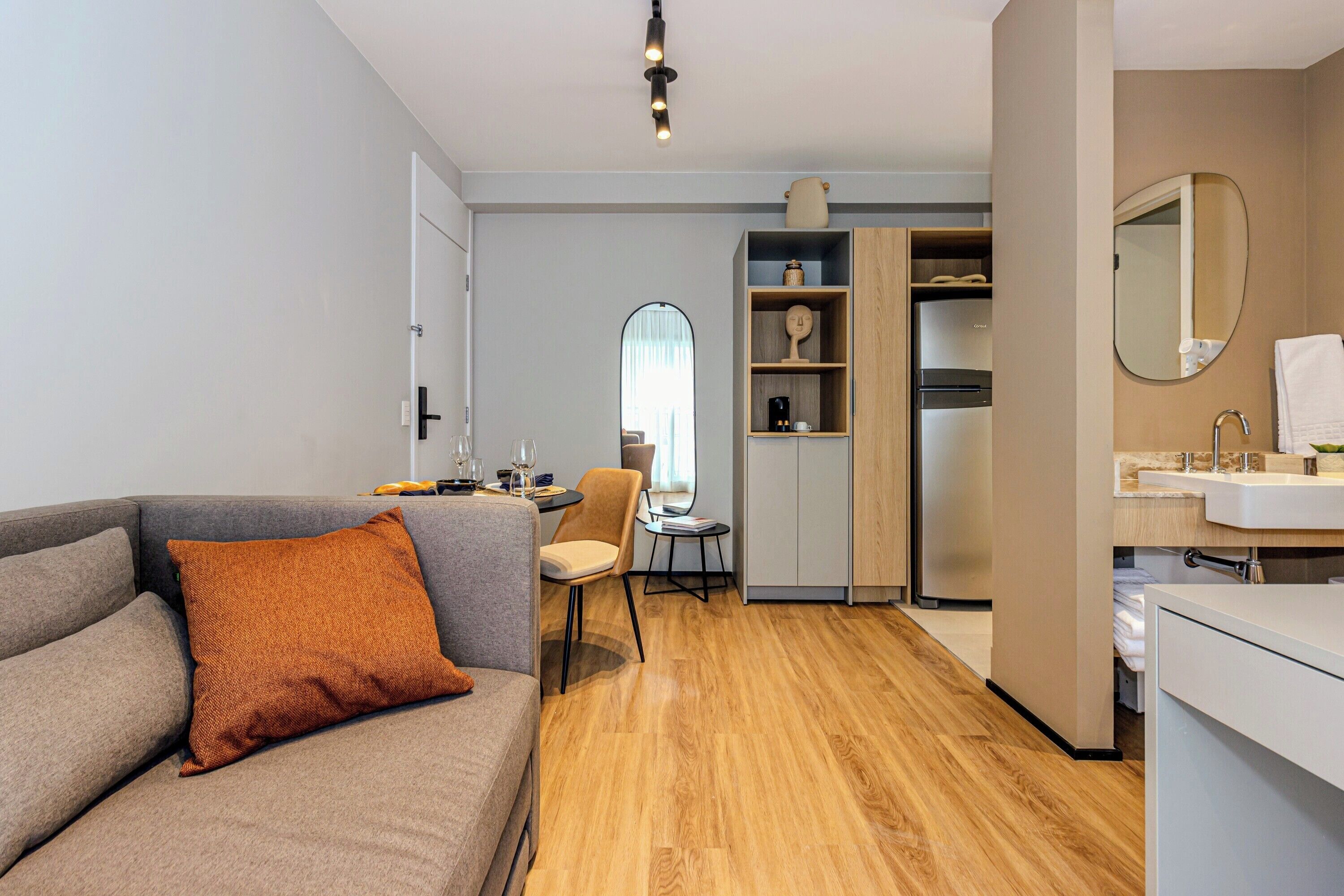 Une chambre d’hôtel moderne avec un canapé gris, un plancher en bois, un coin repas comprenant une chaise et une petite table, ainsi qu’un coin cuisine avec un évier et un miroir.