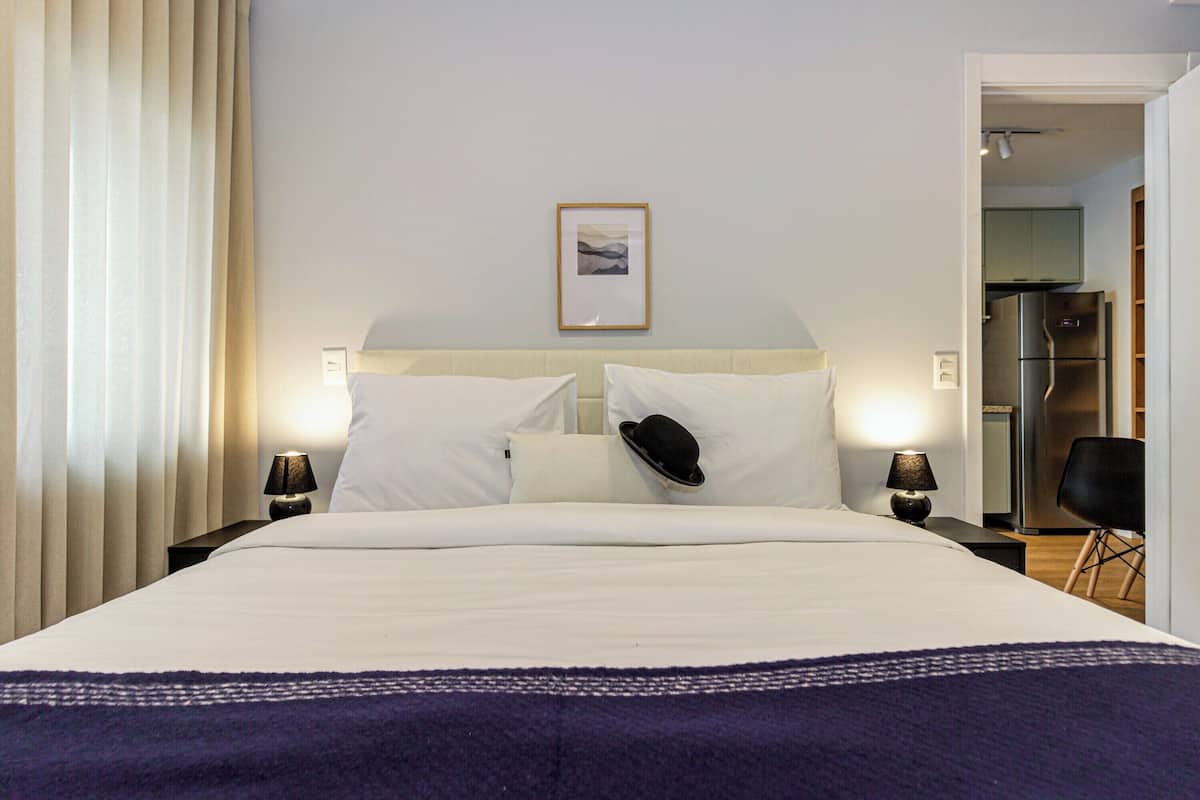 A hotel room with a bed, two bedside tables, a nightstand, a wall-mounted picture, and a view into the kitchen area.