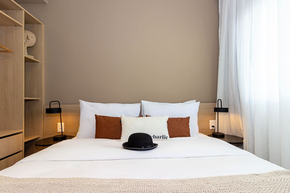 A hotel room with a neatly made bed, a hat on the bed, and a nightstand with a lamp.