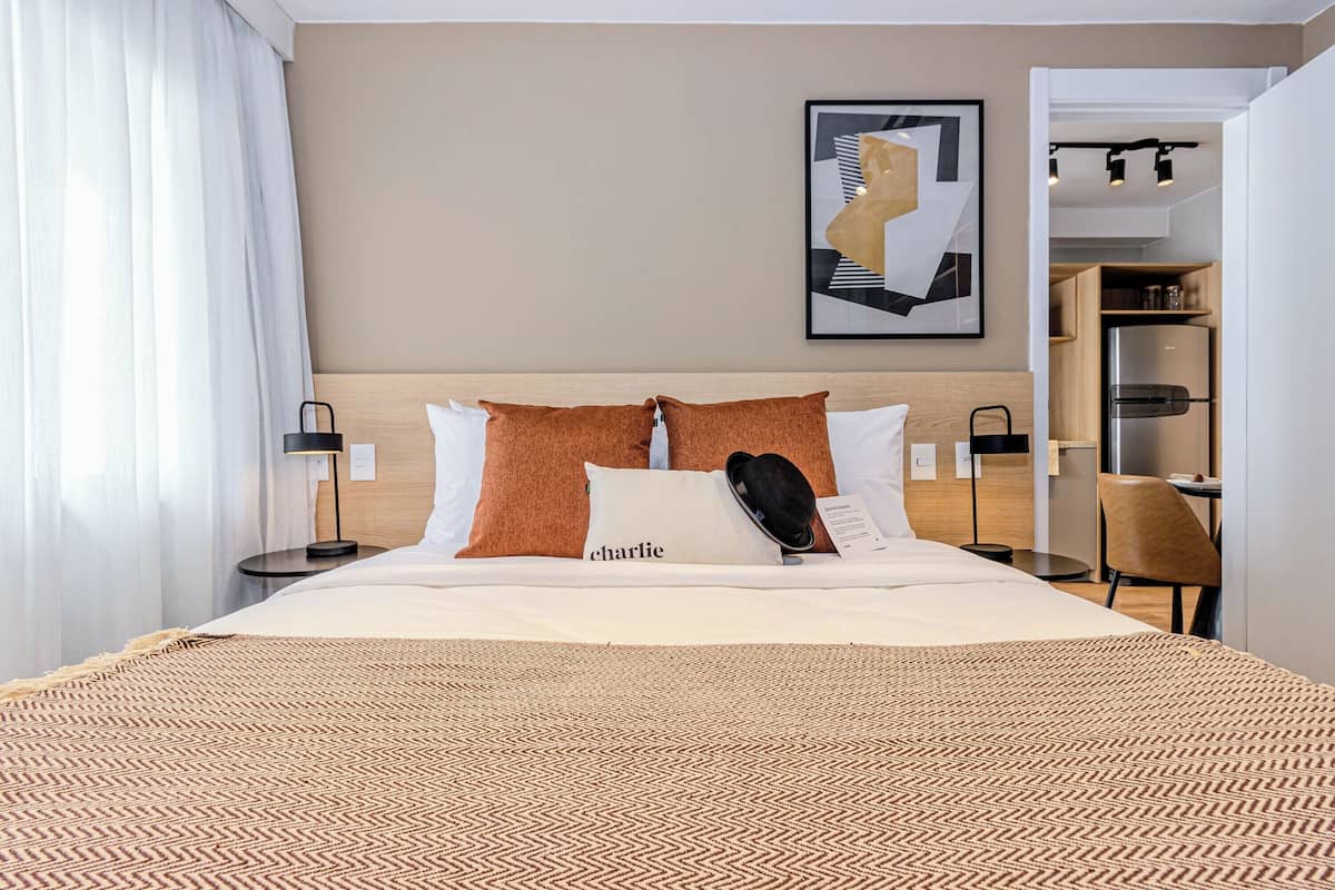 A hotel room with a large bed, a headboard, a framed artwork on the wall, a small table with a chair, and a kitchenette area visible through an open door.
