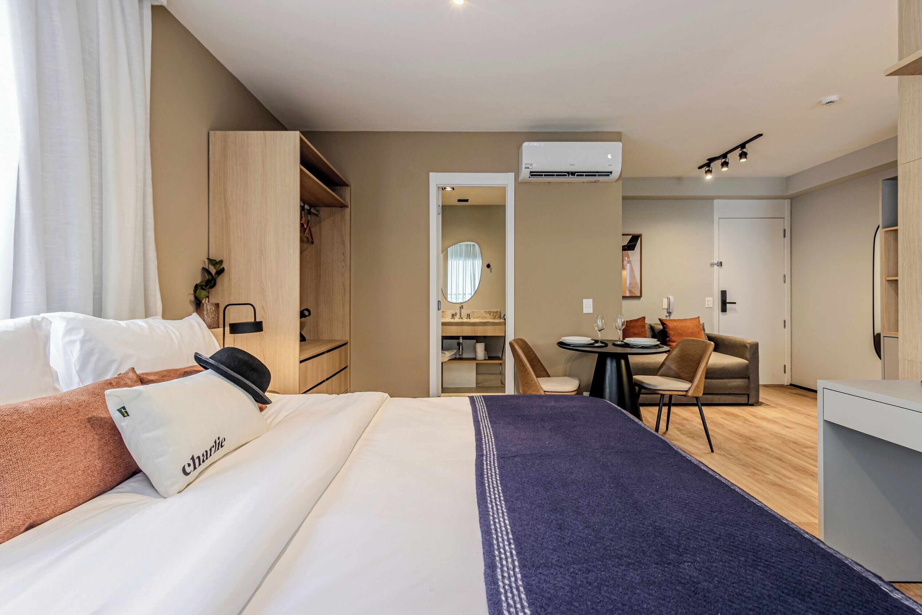 A hotel room with a bed, a desk, a chair, a mirror, and an air conditioning unit.