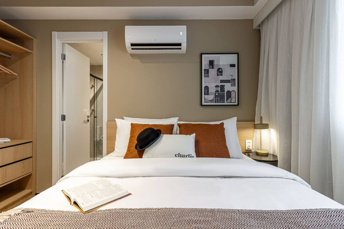 A hotel room with a bed, a bedside table with a lamp, an air conditioning unit, a framed picture on the wall, and a wardrobe.