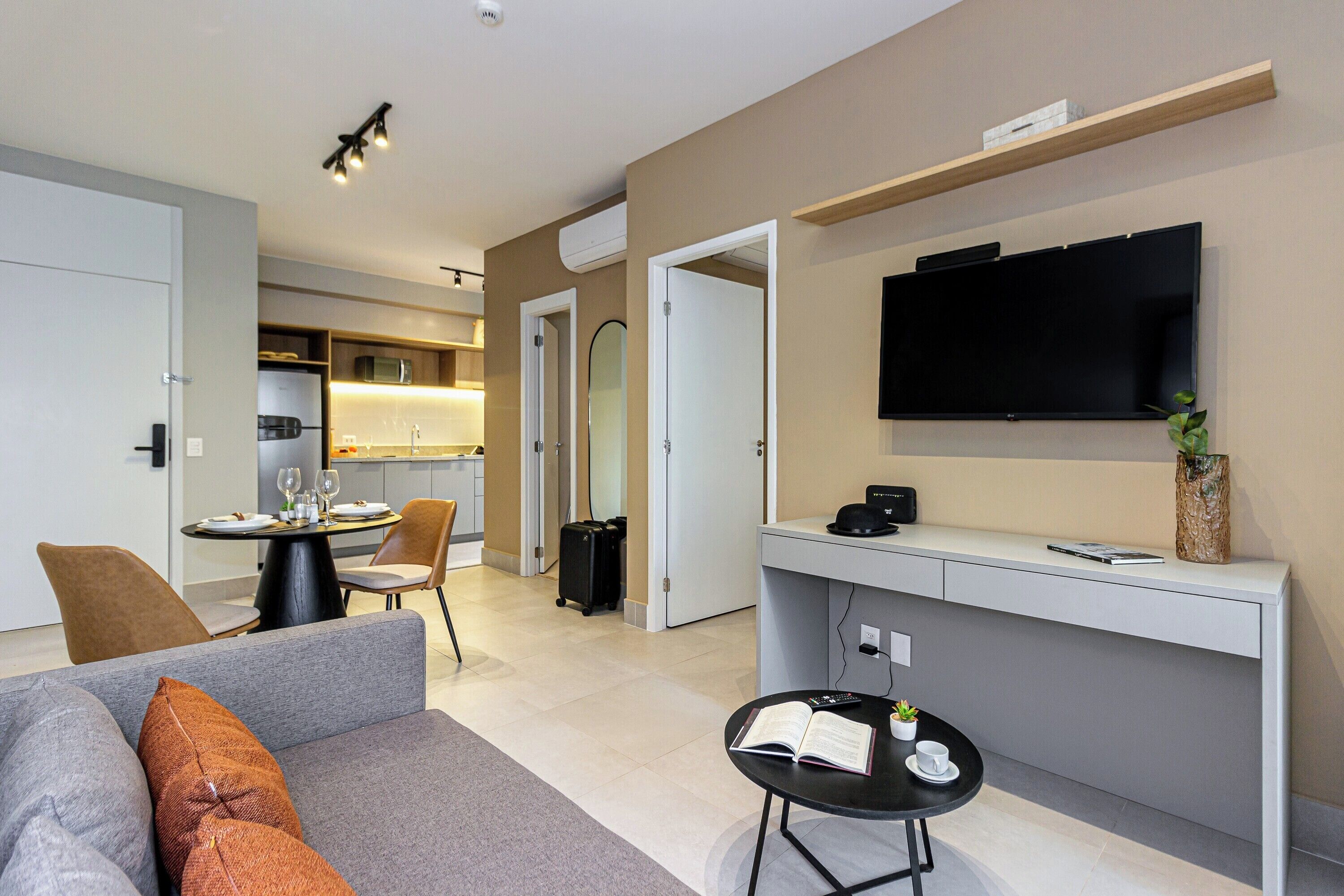 A modern living room with a sofa, a coffee table, a dining area with chairs, a kitchen, and a flat-screen TV mounted on the wall.