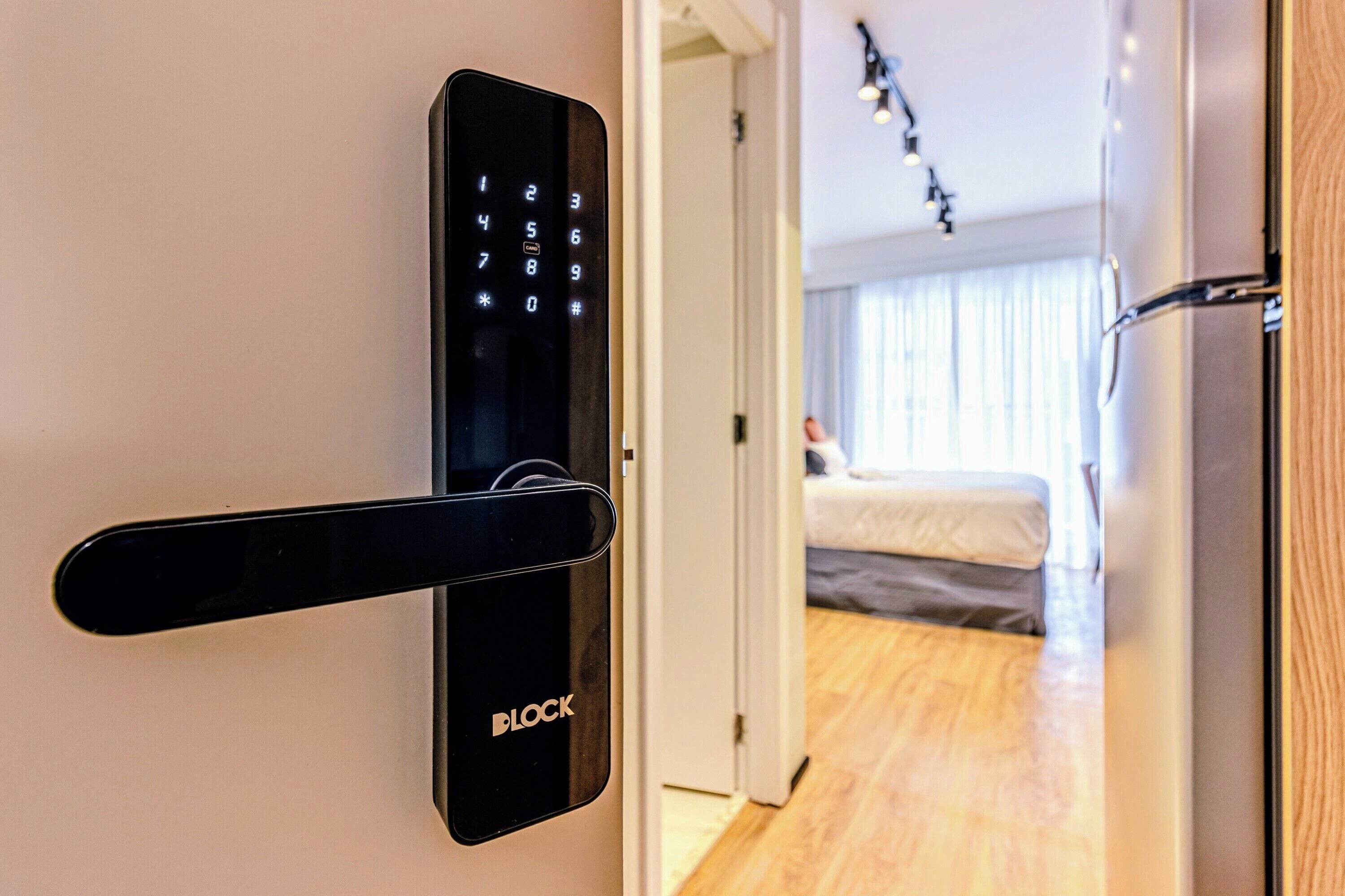 A modern door with a digital keypad lock, a wooden door frame, and a glimpse of a bedroom with a bed and curtains.