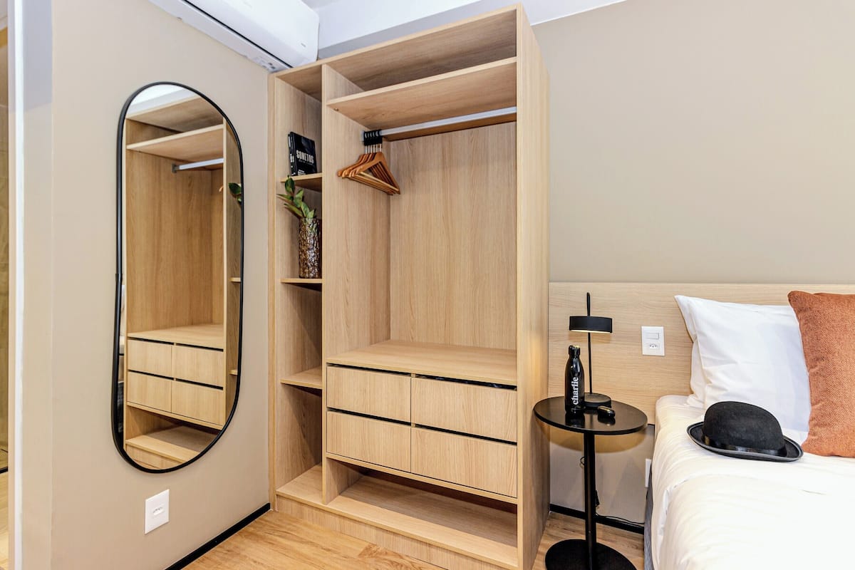 A hotel room with a wooden wardrobe, a mirror, a bed with a white pillow and a black hat, and a small table with a bottle and a plant.