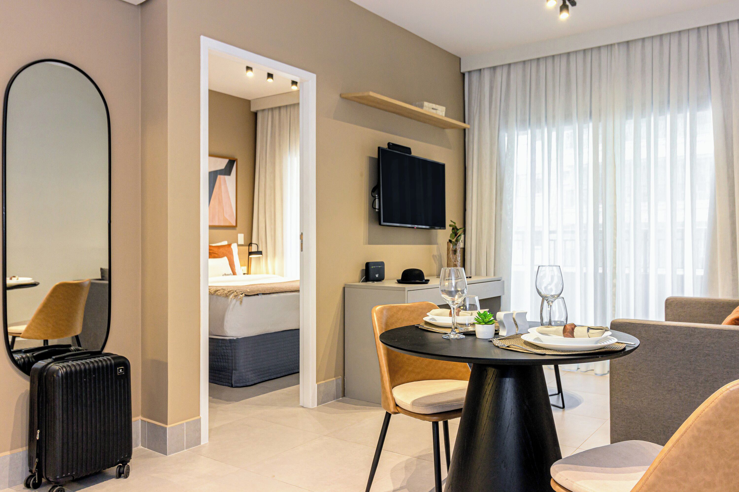 A hotel room with a dining area, a TV, a mirror, and a suitcase.