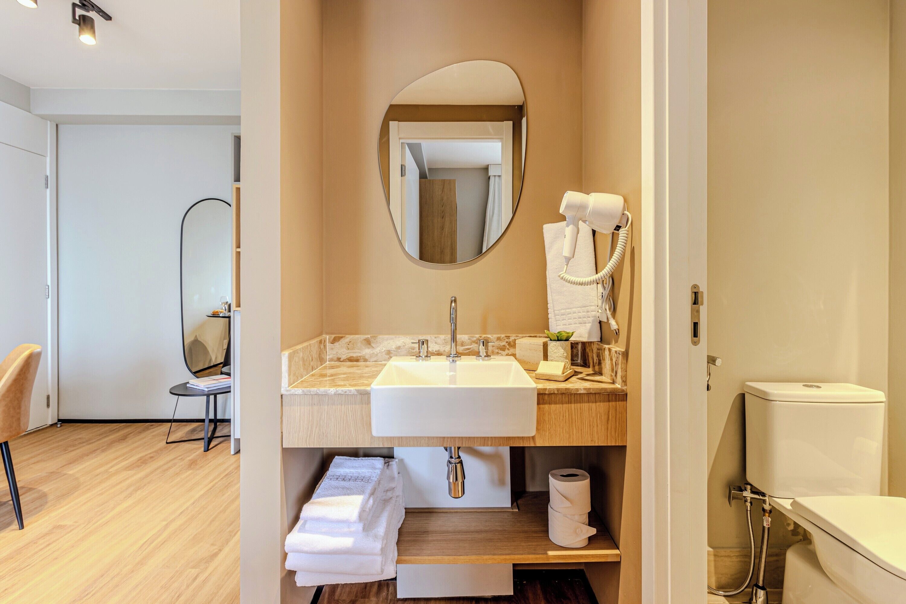A modern bathroom with a sink, mirror, and toilet. There is a towel rack with towels and a hairdryer. The room has wooden flooring and a mirror on the wall.