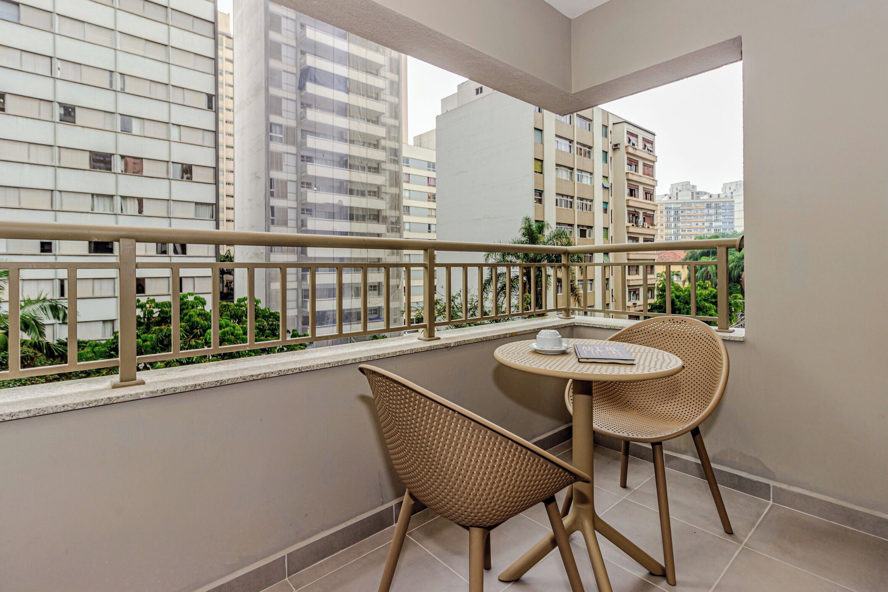 A balcony with a round table and two wicker chairs, overlooking a cityscape with tall buildings.