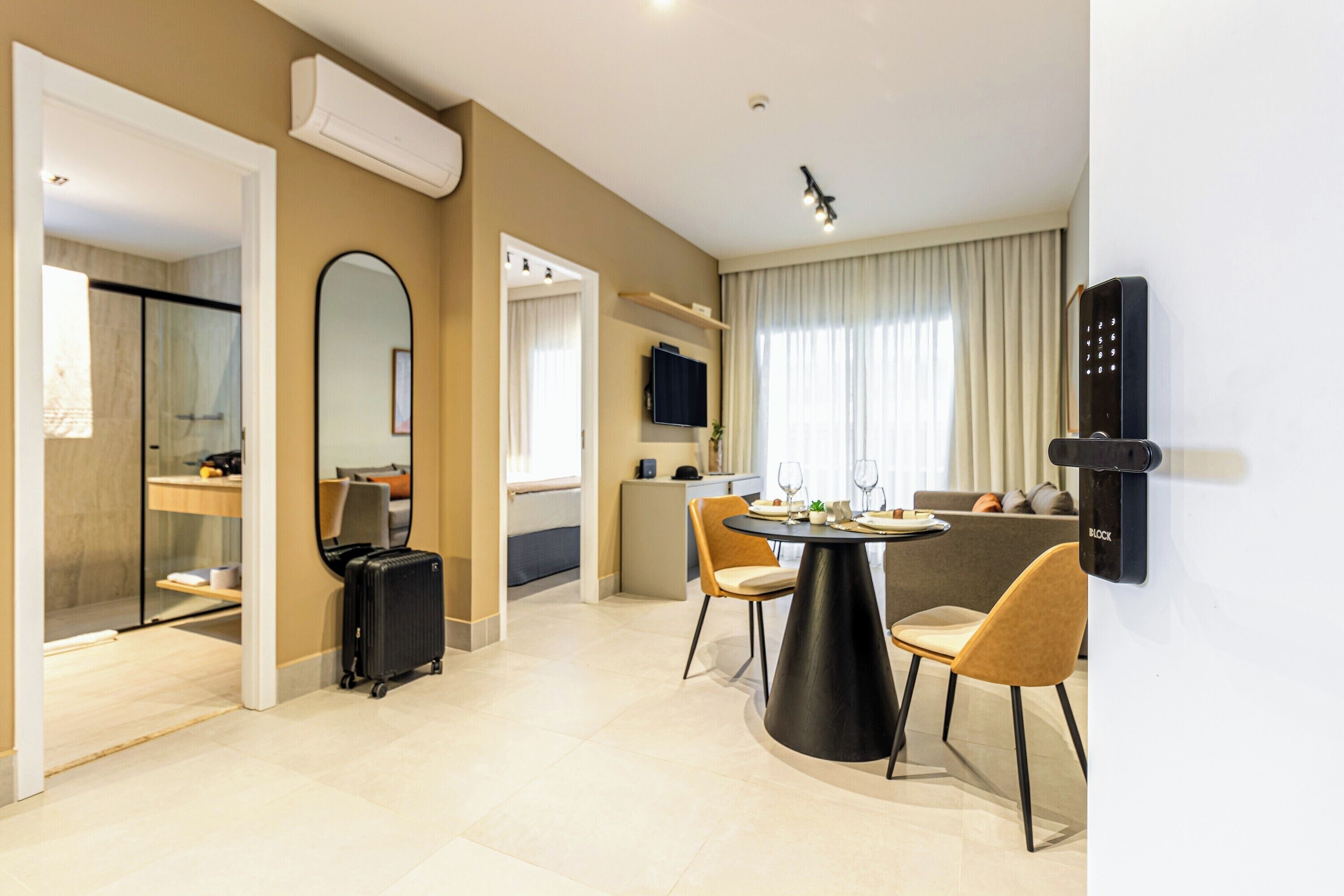 A modern hotel room with a dining area, a large mirror, a black suitcase, and a wall-mounted control panel.