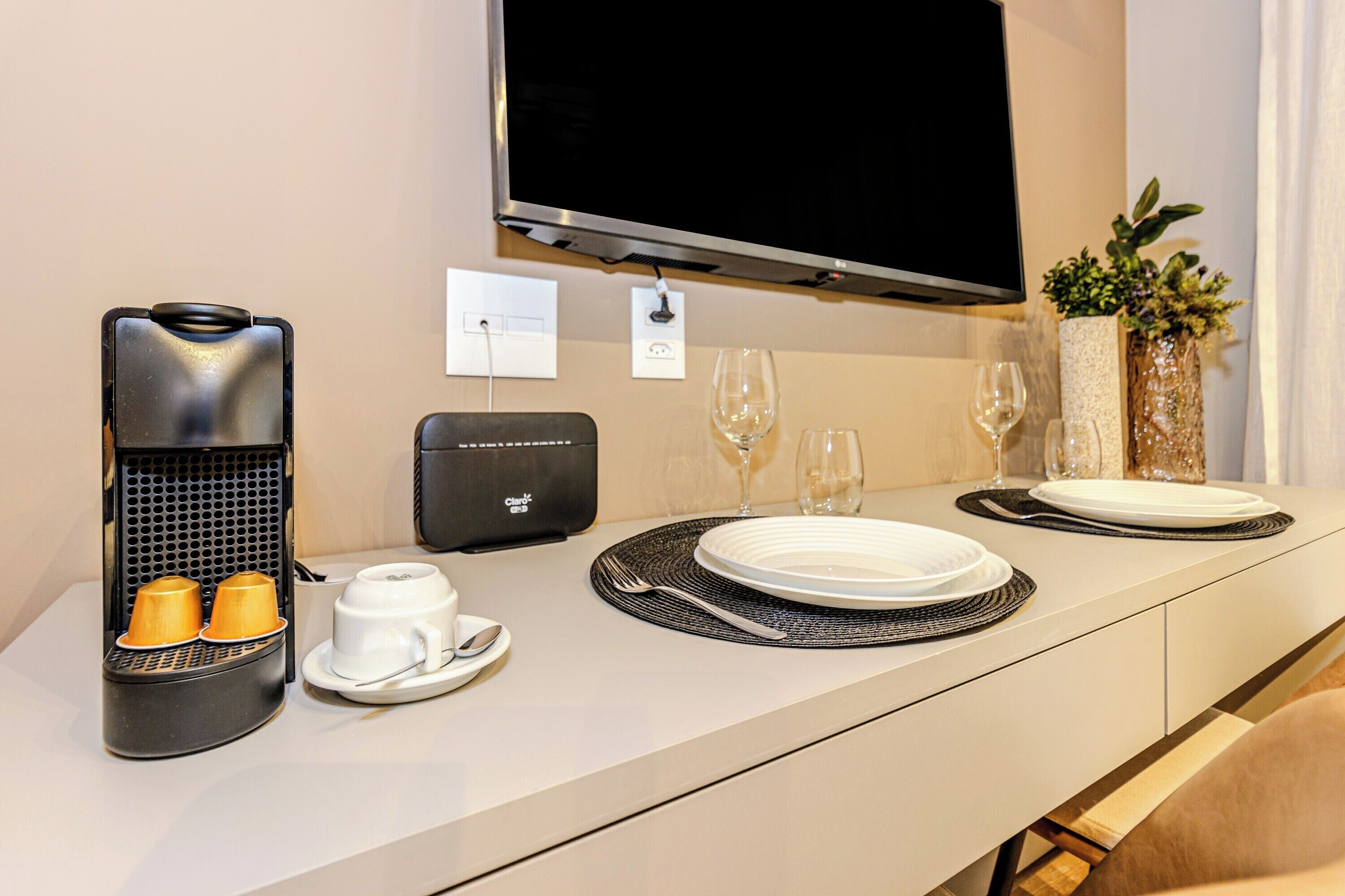 A hotel room with a modern kitchenette, a flat-screen TV mounted on the wall, a small potted plant, and a neatly arranged dining table with plates, glasses, and a coffee maker.