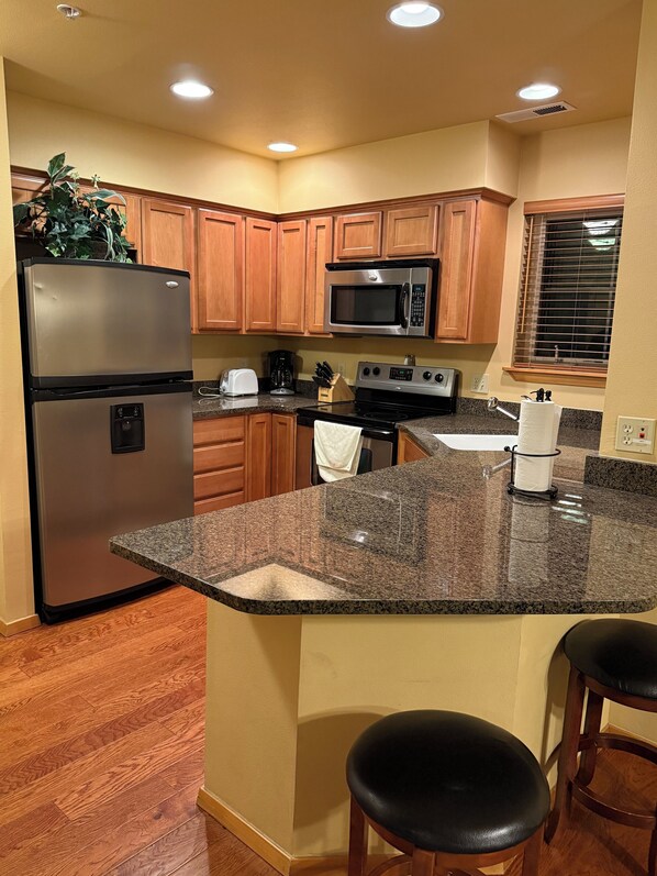 A kitchen with wooden cabinets, a stainless steel refrigerator, a microwave, a stove, granite countertops, and wooden bar stools.