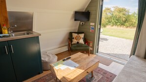 Une petite cuisine équipée comprenant un évier, une chaise agrémentée d’un coussin décoratif, une petite table en bois sur laquelle se trouvent des papiers, et une vue sur un espace verdoyant à travers une porte vitrée.