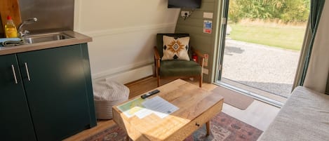 Une petite cuisine équipée comprenant un évier, une chaise agrémentée d’un coussin décoratif, une petite table en bois sur laquelle se trouvent des papiers, et une vue sur un espace verdoyant à travers une porte vitrée.