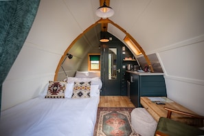 Une chambre mansardée chaleureuse, avec un plafond en pente, un lit agrémenté d’oreillers à motifs, un petit bureau et une vue sur la cuisine.