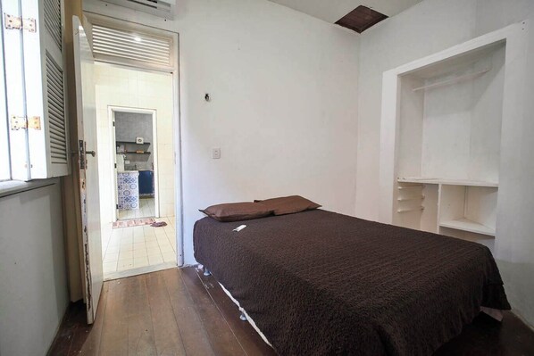 A small, sparsely furnished room with a bed, a door leading to a bathroom, and a white wall with a shelf unit. - Casa com Piscina Churrasqueira e Wi-fi Cumbuco (Caucaia)