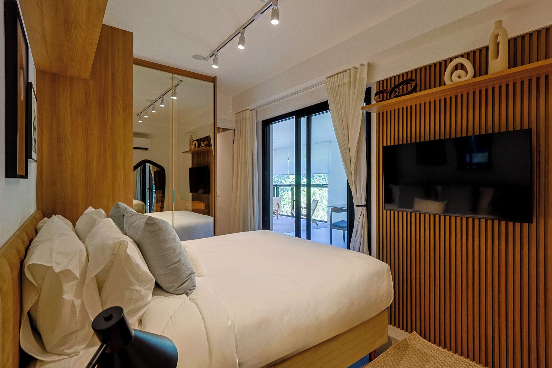 Een slaapkamer met een groot bed, een televisie bevestigd aan een houten paneel, en een balkon met uitzicht.
