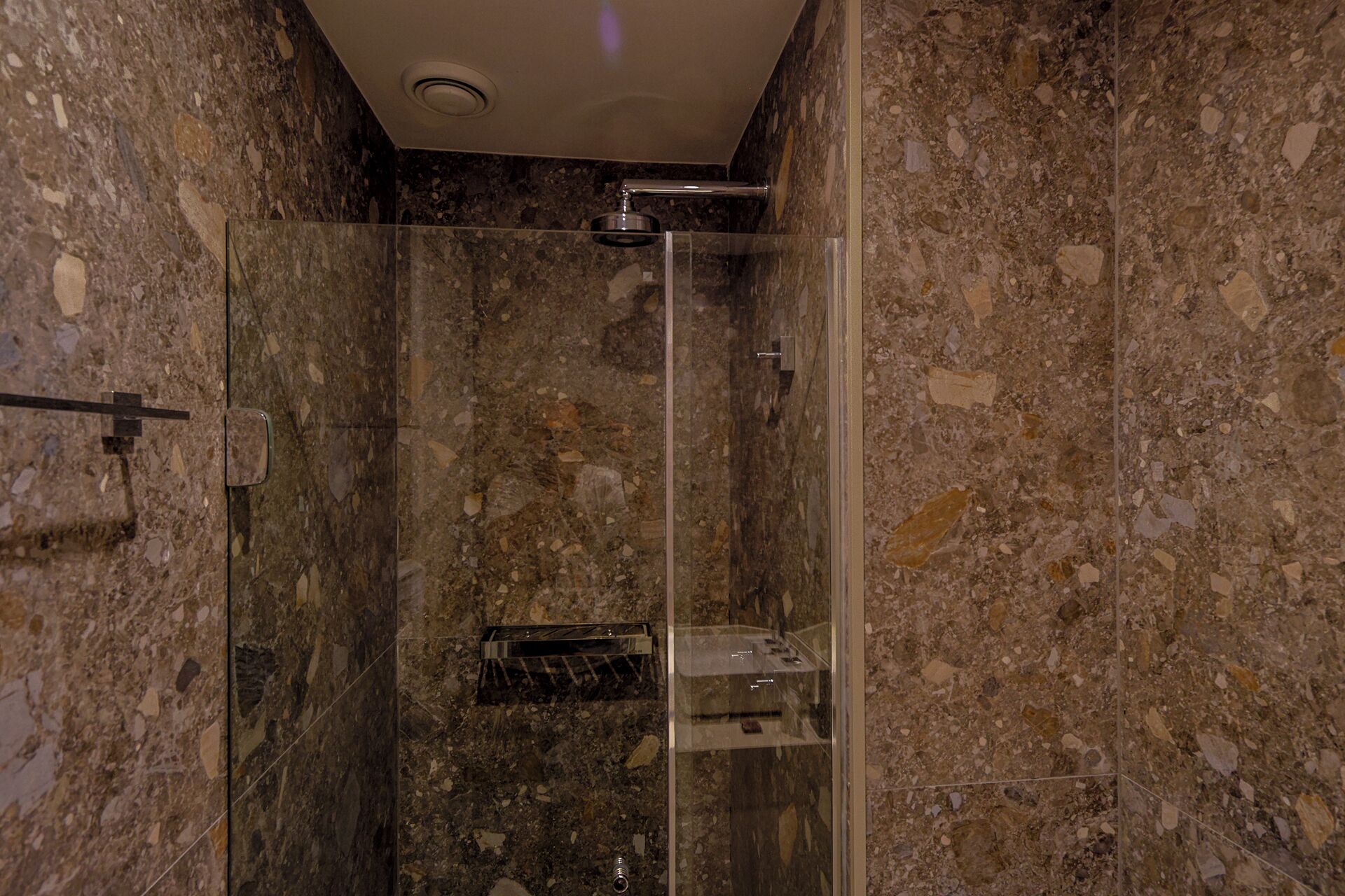 A glass-enclosed shower stall with a marble wall and a ceiling vent.