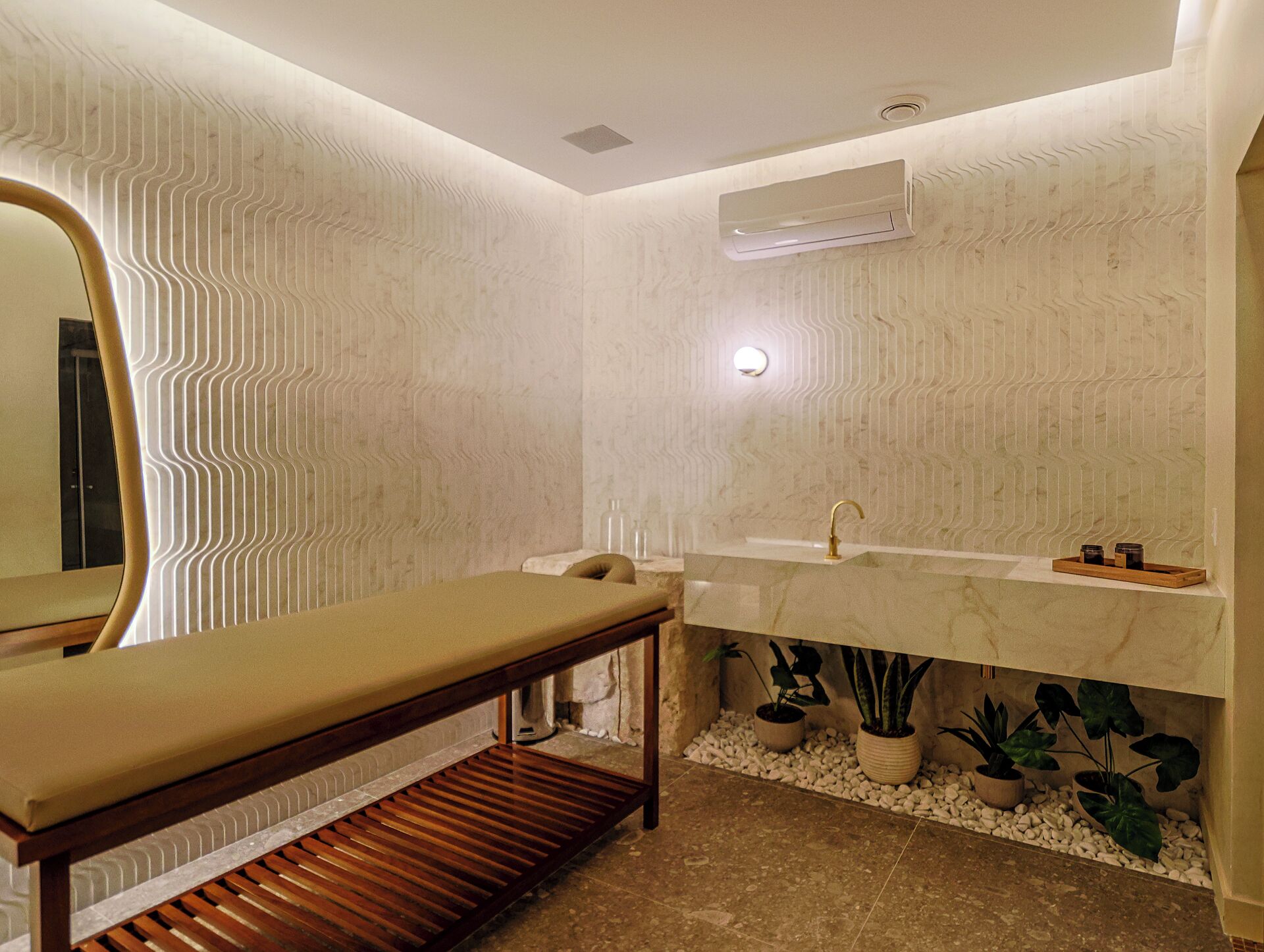 A modern bathroom with a wooden bench, a sink with a gold faucet, and a mirror with a unique wavy pattern.