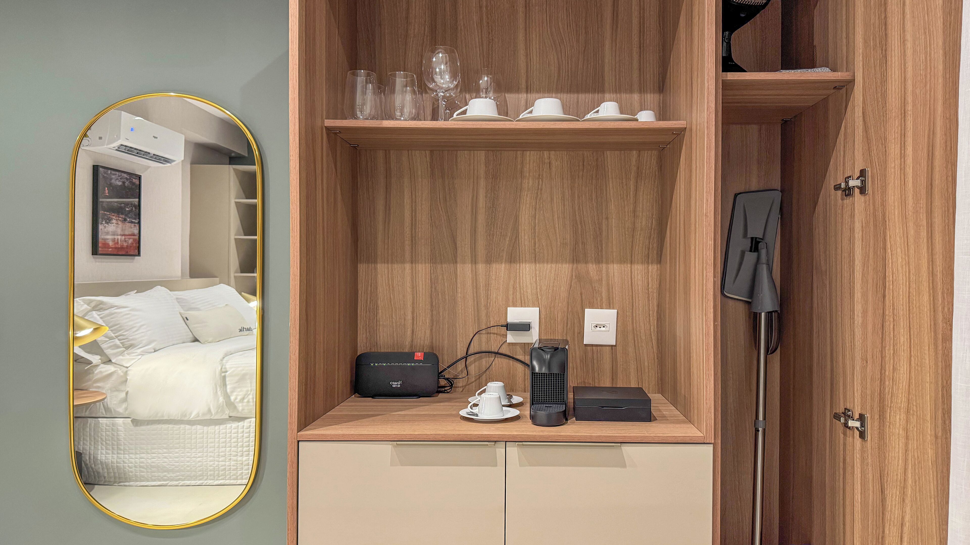 Une petite pièce comprenant un lit, un miroir, une étagère en bois avec des tasses et des verres, une machine à café et un aspirateur.