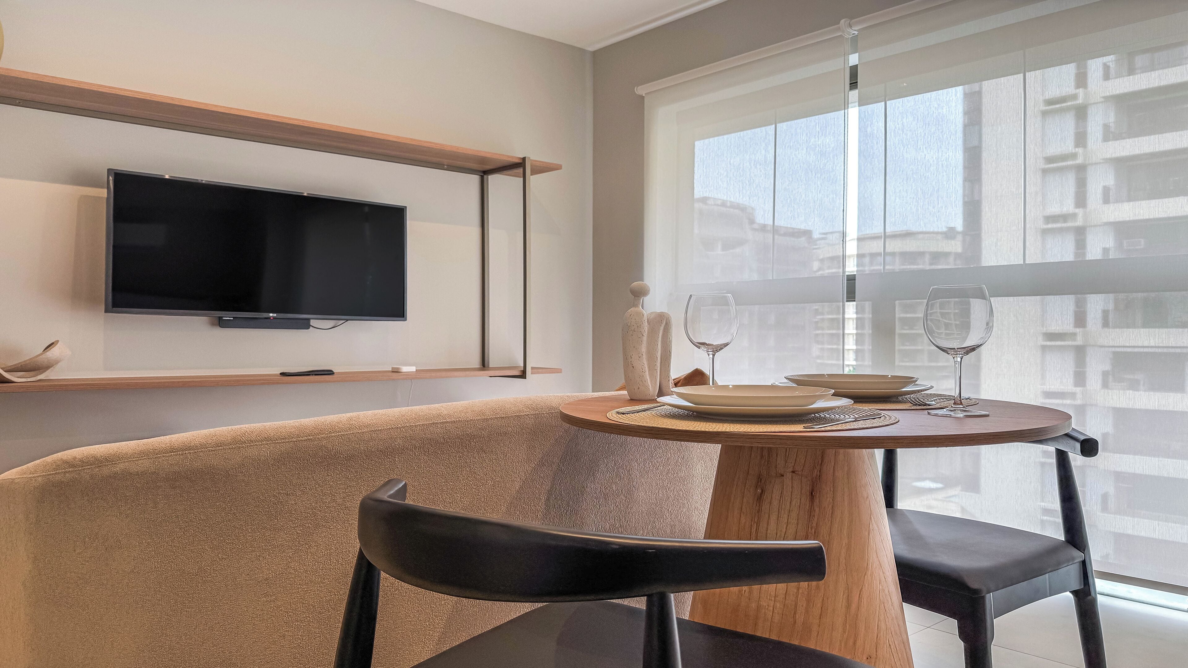 Un comedor moderno con una mesa redonda, dos sillas, un televisor montado en la pared y un ventanal con vistas a edificios.