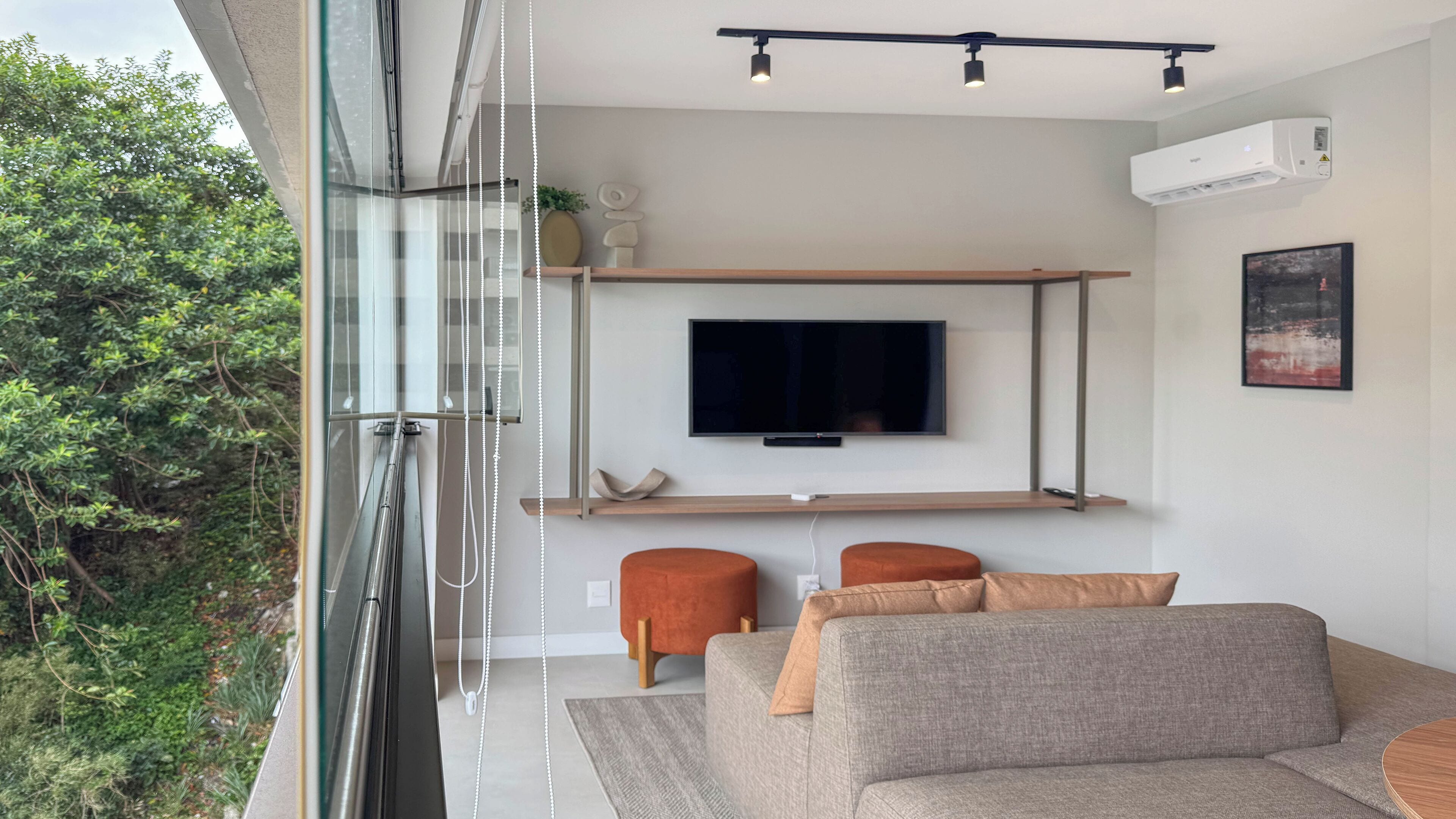 A modern living room with a large flat-screen TV mounted on a wooden shelf, a grey sofa with orange cushions, and a glass wall overlooking greenery.